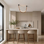 One lit Clérachic loop pendant in black and brown hangs over a kitchen island with three bar stools beside a tall window.