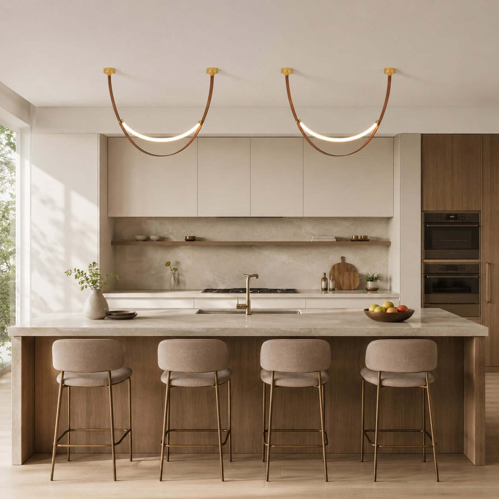 Two gold-capped Clérachic loop pendants hang side by side in a pale kitchen above a stone island and four stools.