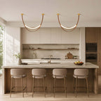 Two gold-capped Clérachic loop pendants hang side by side in a pale kitchen above a stone island and four stools.
