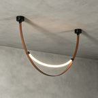 A black-capped Clérachic loop pendant, lit, against a rough concrete ceiling, with its brown strap curving beneath the LED strip.