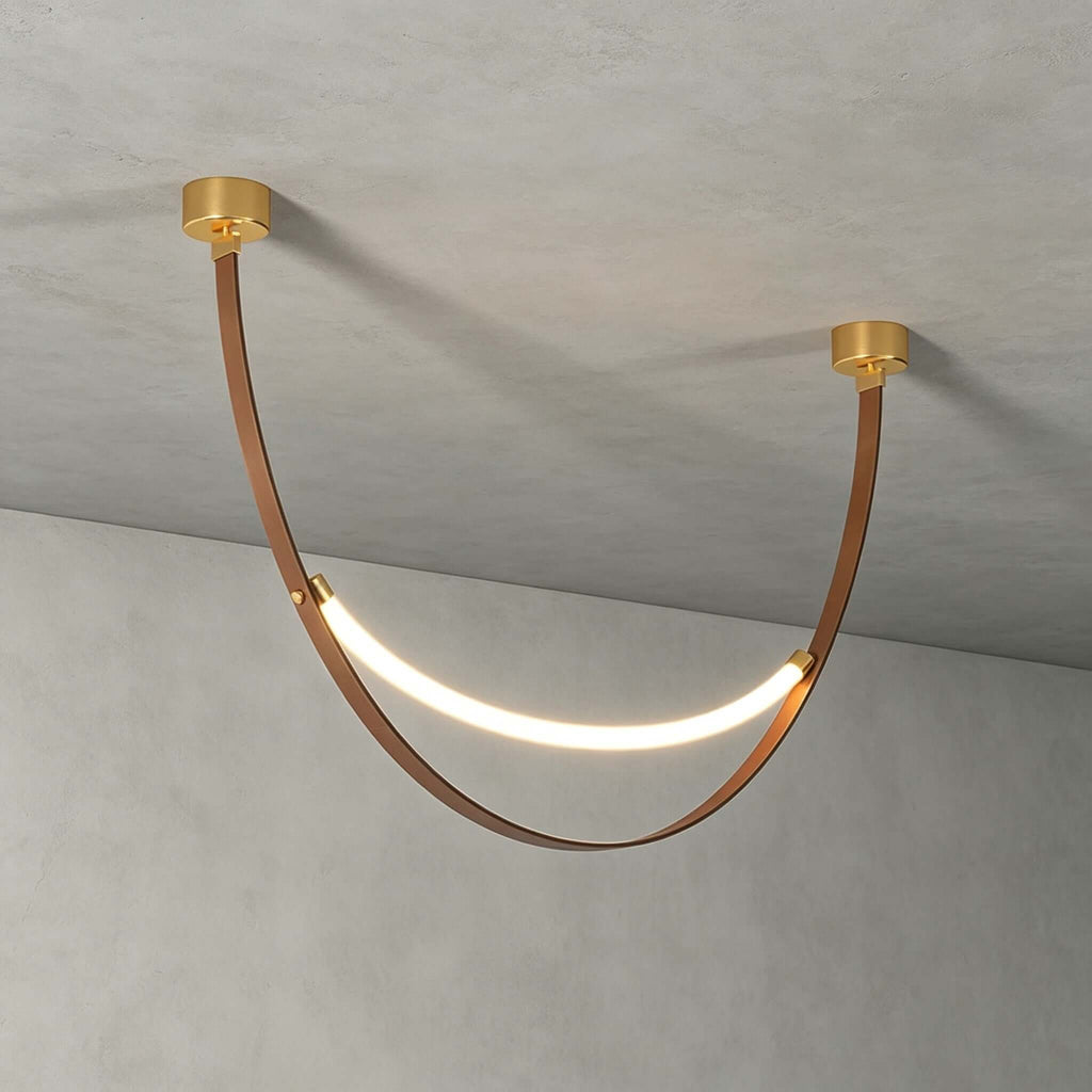 A single gold-capped Clérachic loop pendant, lit, hanging against a grey concrete ceiling and wall, its brown strap in a deep curve.