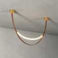 A single gold-capped Clérachic loop pendant, lit, hanging against a grey concrete ceiling and wall, its brown strap in a deep curve.