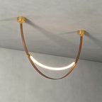 A single gold-capped Clérachic loop pendant, lit, hanging against a grey concrete ceiling and wall, its brown strap in a deep curve.