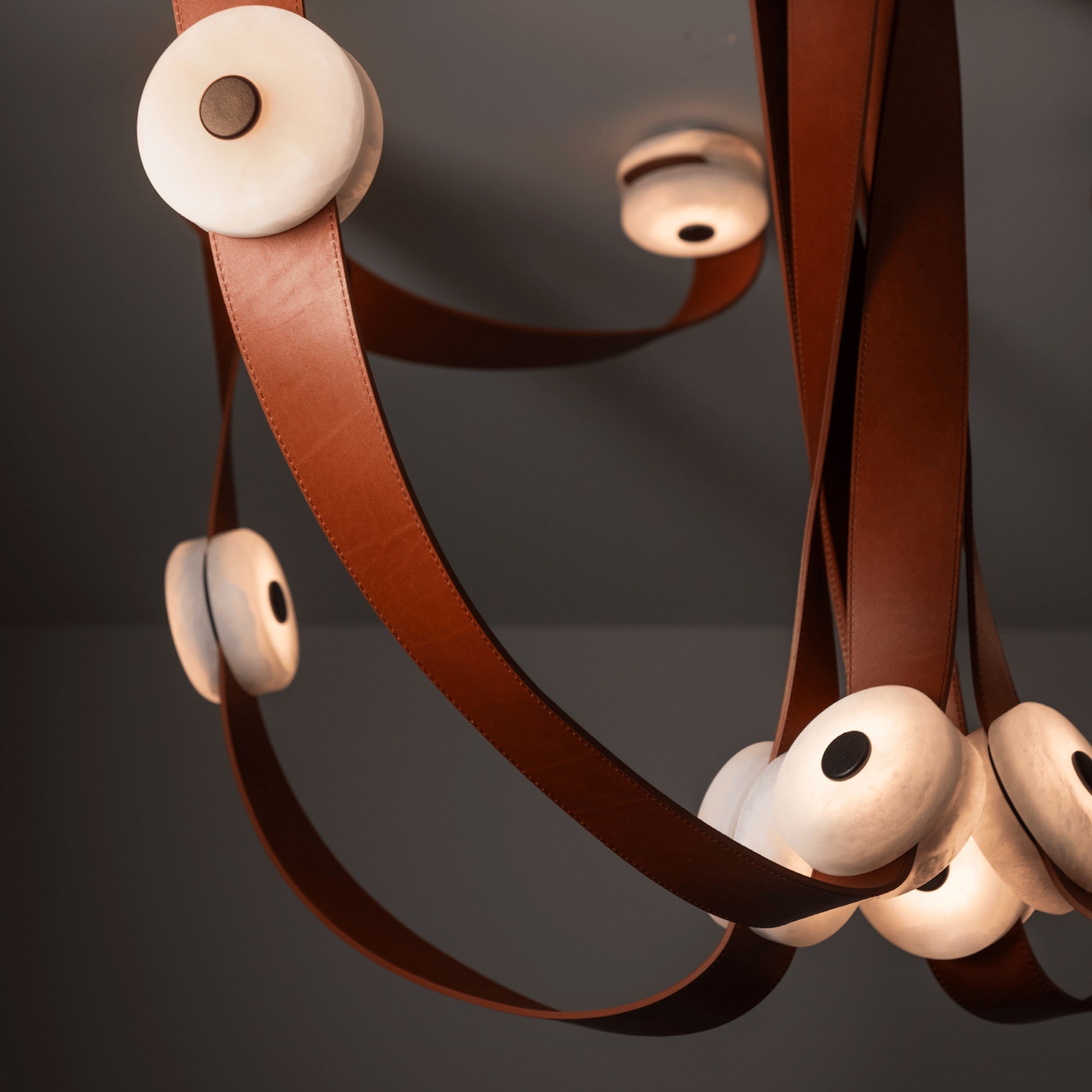 Clérachic Leather Strap Chandelier close up, cognac leather curving past lit alabaster discs on a dark ceiling.