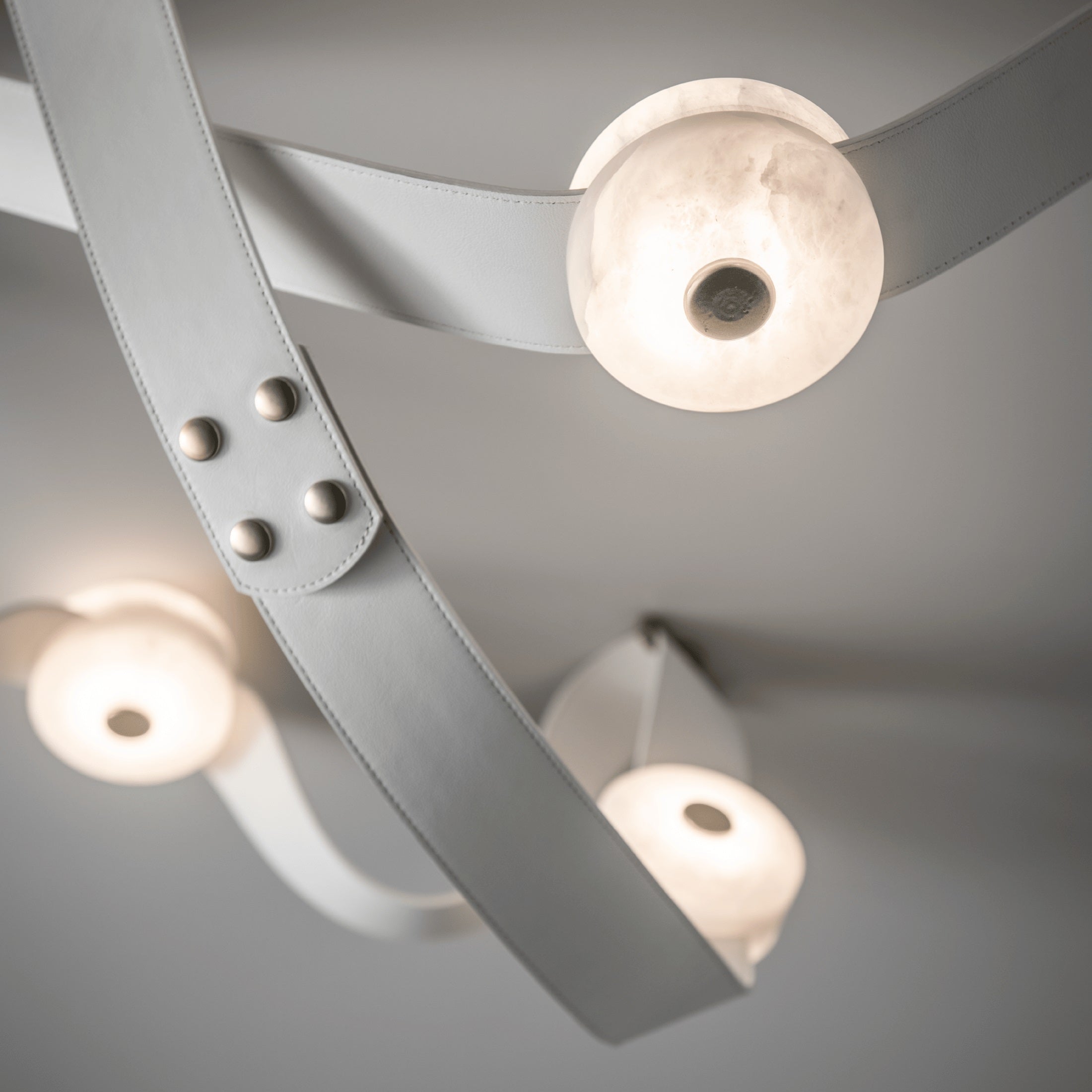 Close-up of steel studs and pale leather on the Clérachic Leather Strap Chandelier beside a glowing alabaster disc.