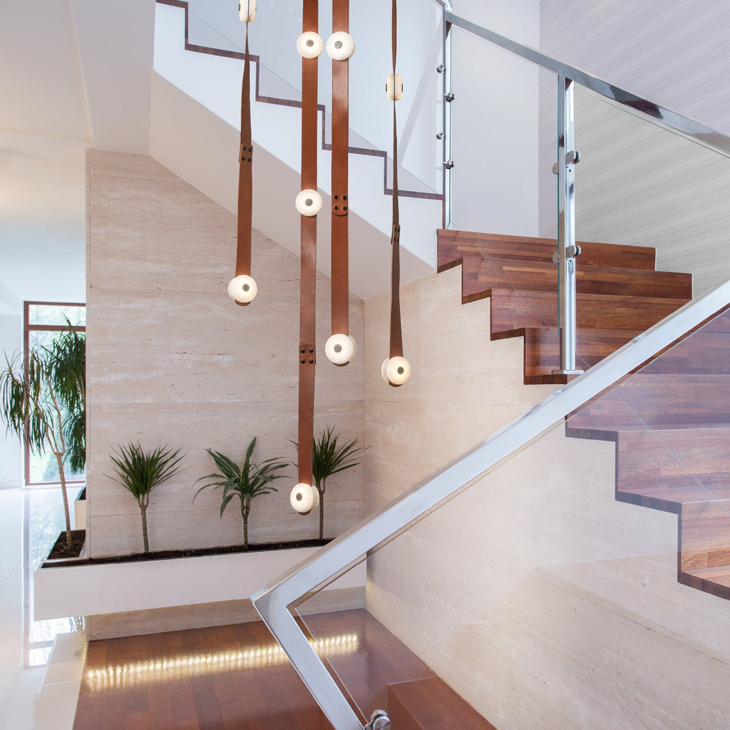 Cognac Clérachic Leather Strap Chandelier falling down a travertine stairwell beside a glass and steel balustrade.