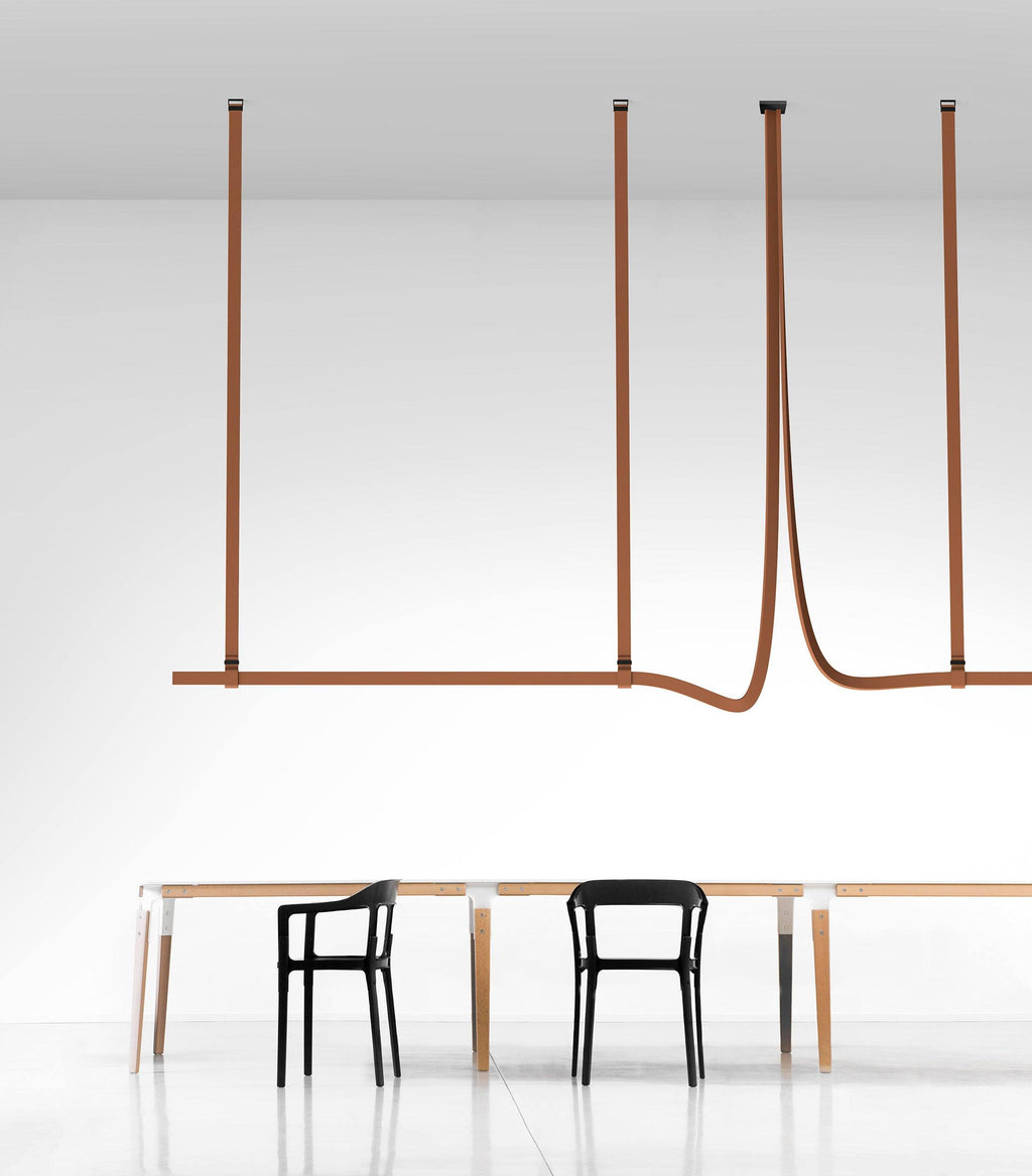 Tan leather straps suspend a slim horizontal light bar above a long table with black chairs, a Clérachic strap pendant in a white room.