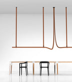 Tan leather straps suspend a slim horizontal light bar above a long table with black chairs, a Clérachic strap pendant in a white room.
