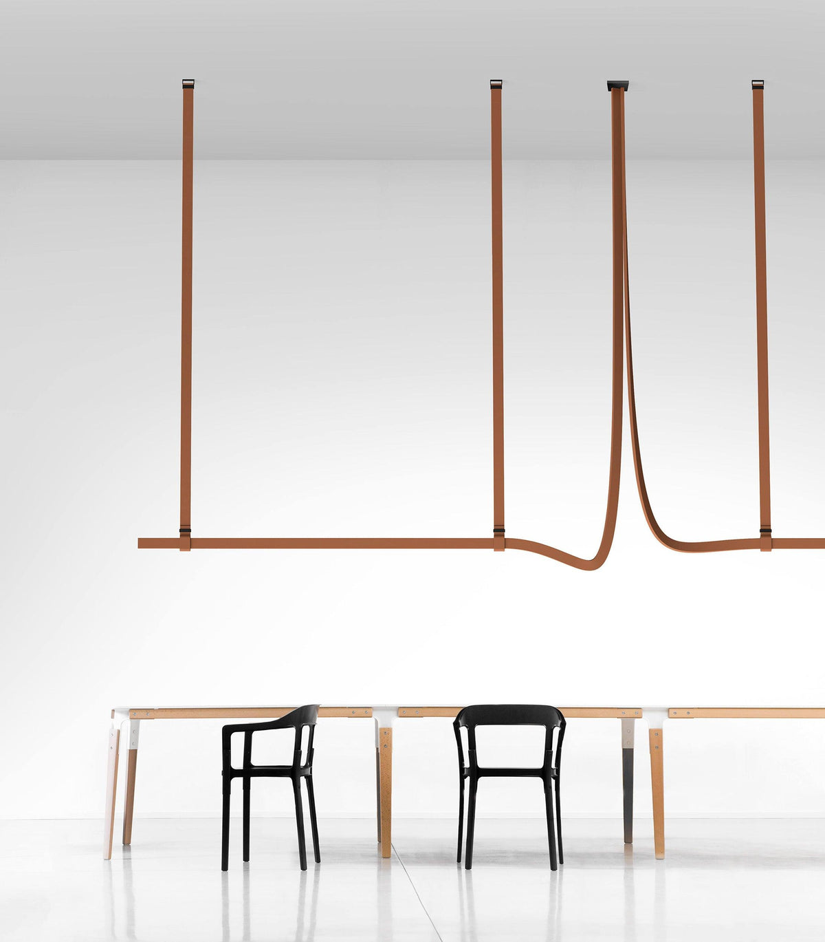 Tan leather straps suspend a slim horizontal light bar above a long table with black chairs, a Clérachic strap pendant in a white room.