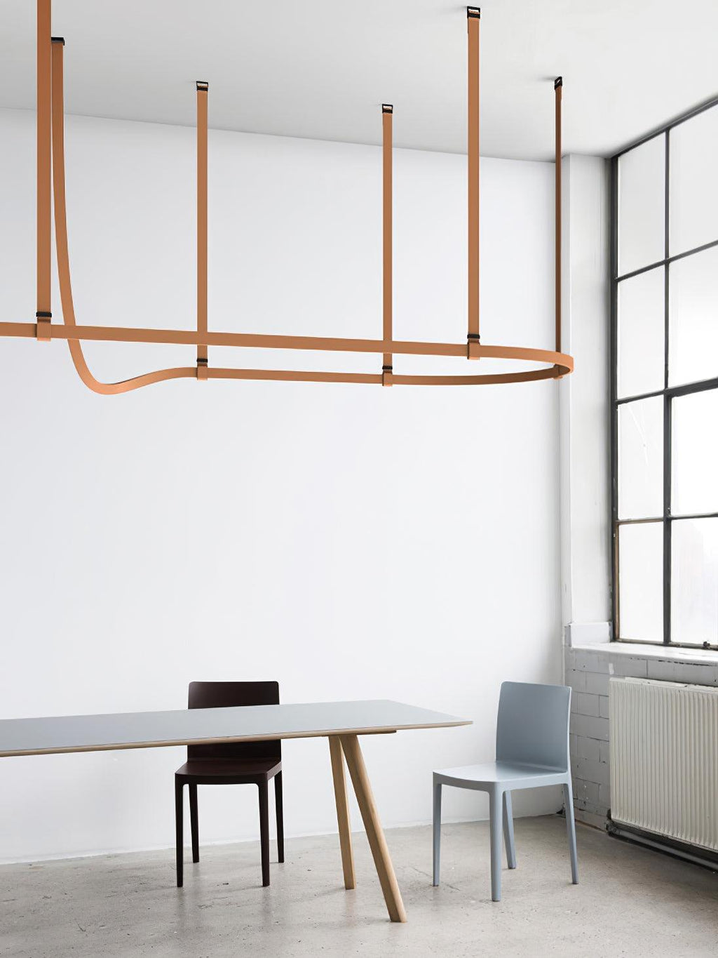 This Clérachic strap pendant hangs a horizontal light bar from four tan leather straps beside a tall steel-framed window.
