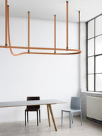 This Clérachic strap pendant hangs a horizontal light bar from four tan leather straps beside a tall steel-framed window.