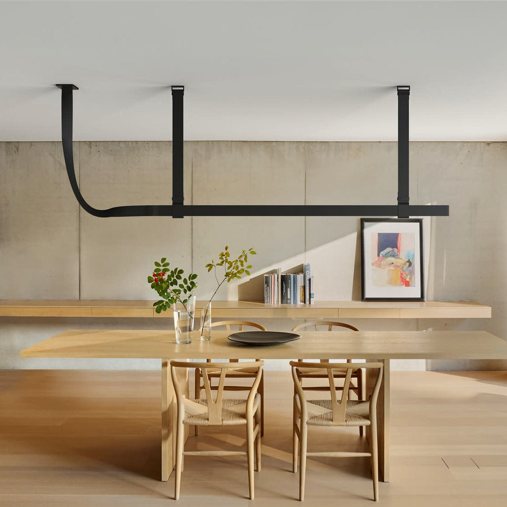 Black leather strap pendant carrying a long light bar above a pale wood dining table, a wooden bench and a framed print, from Clérachic.