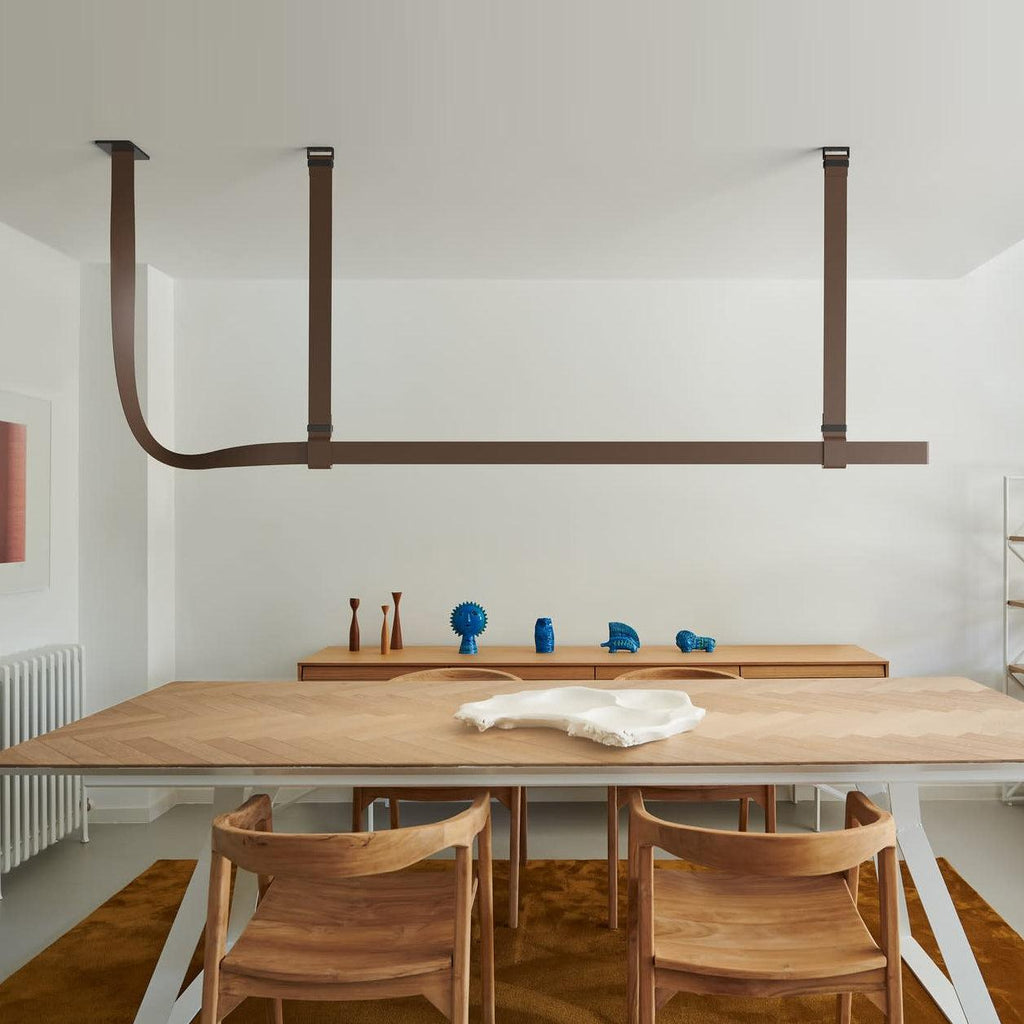 Brown leather straps hold a Clérachic light bar above a herringbone wood dining table and wooden chairs in a white room.