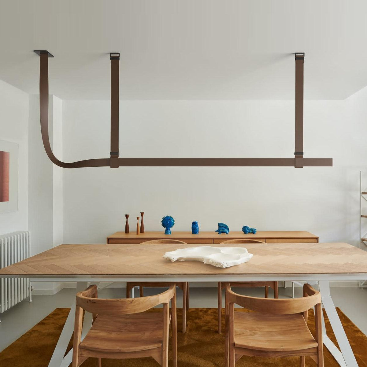 Brown leather straps hold a Clérachic light bar above a herringbone wood dining table and wooden chairs in a white room.