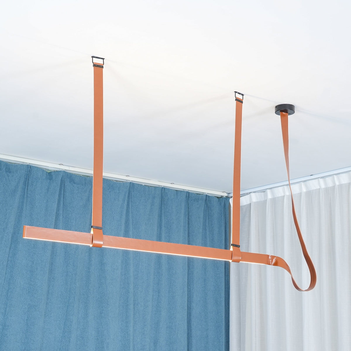 Tan leather straps and a slim light bar hang below a ceiling canopy, with blue and white curtains behind, from Clérachic.