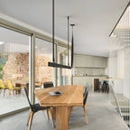 Black strap pendant above a solid wood dining table in a glass-walled room with a stone wall, from Clérachic.