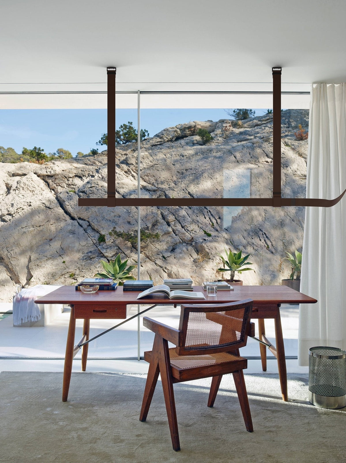 Brown strap pendant hung in front of a glazed wall with a rocky view, above a wooden desk and cane chair, from Clérachic.