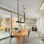 Black strap pendant above a wood dining table beside a glass wall, with yellow chairs and a stone wall beyond, from Clérachic.