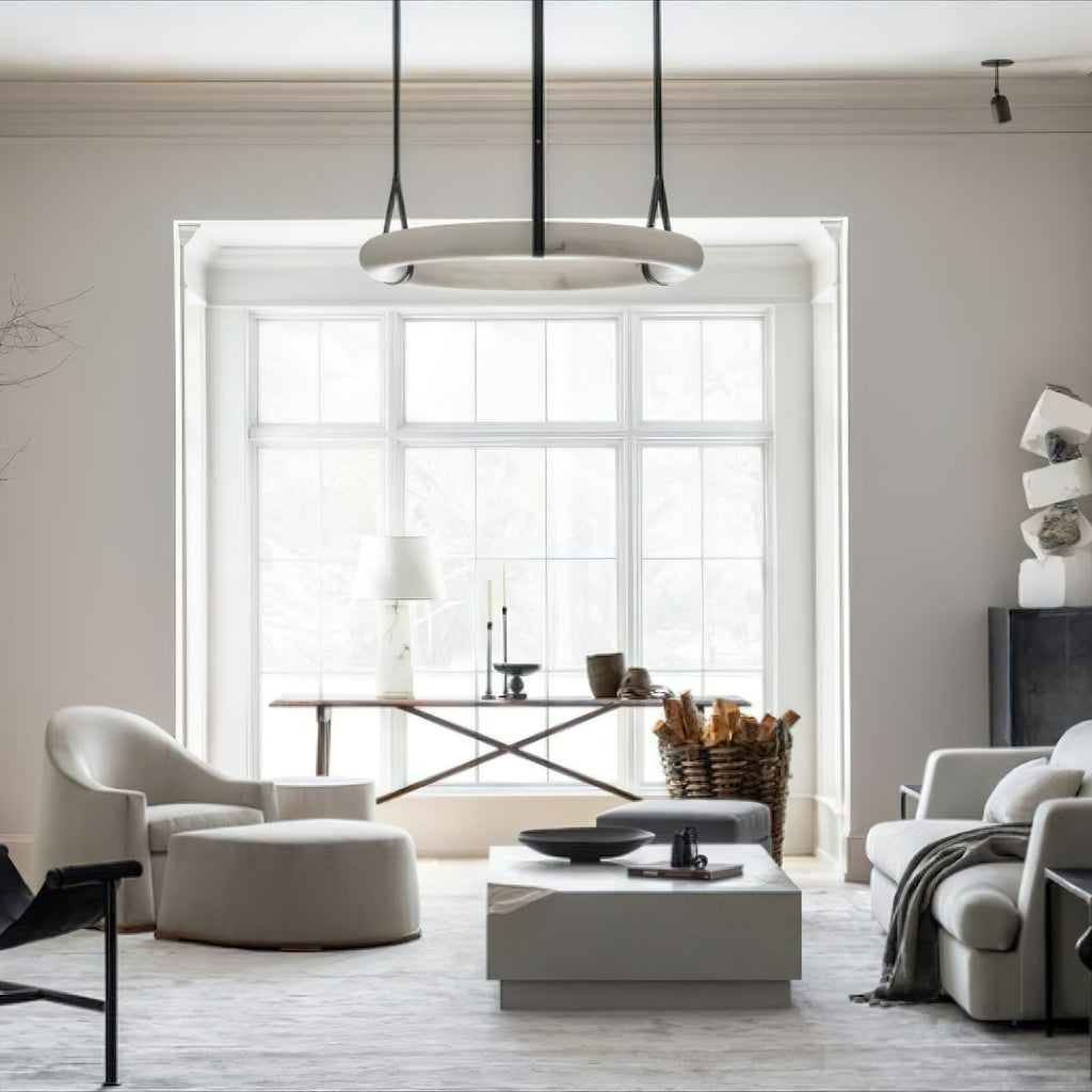 A Clérachic alabaster ring chandelier suspended in a bright bay window above a white living room with linen armchairs.