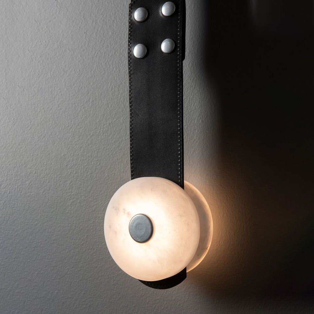 Black leather and nickel rivets on the Clérachic Leather Strap Wall Sconce, alabaster disc glowing low on the strap.