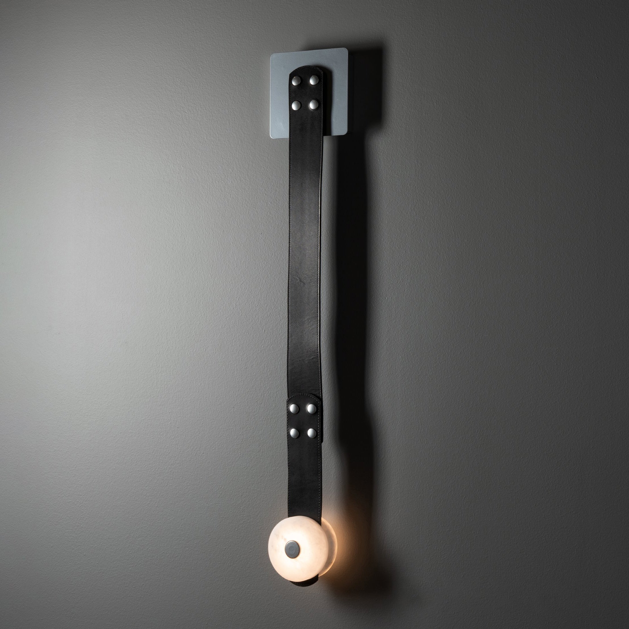 Clérachic Leather Strap Wall Sconce in its long one-head size, black strap on a brushed steel plate above a lit disc.