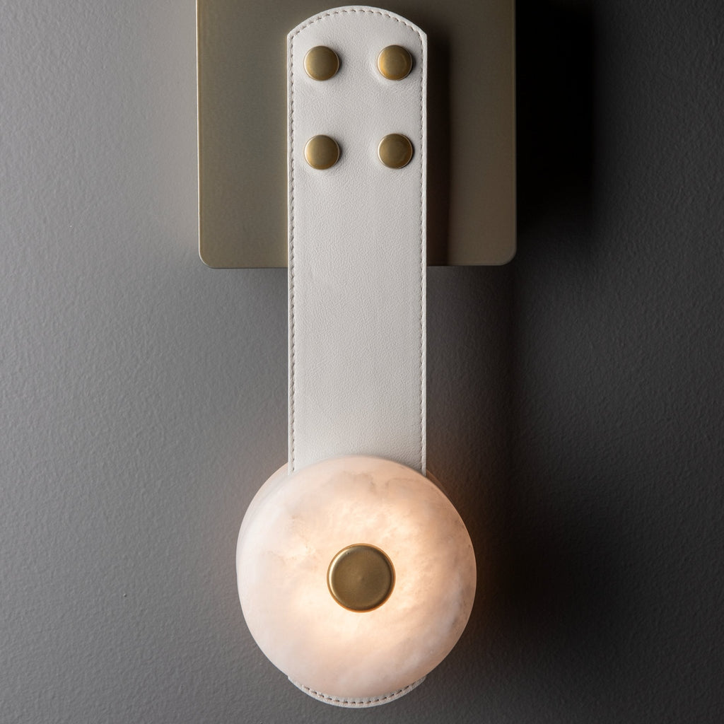 Close view of the Clérachic Leather Strap Wall Sconce in white leather, brass rivets above a softly lit alabaster disc.