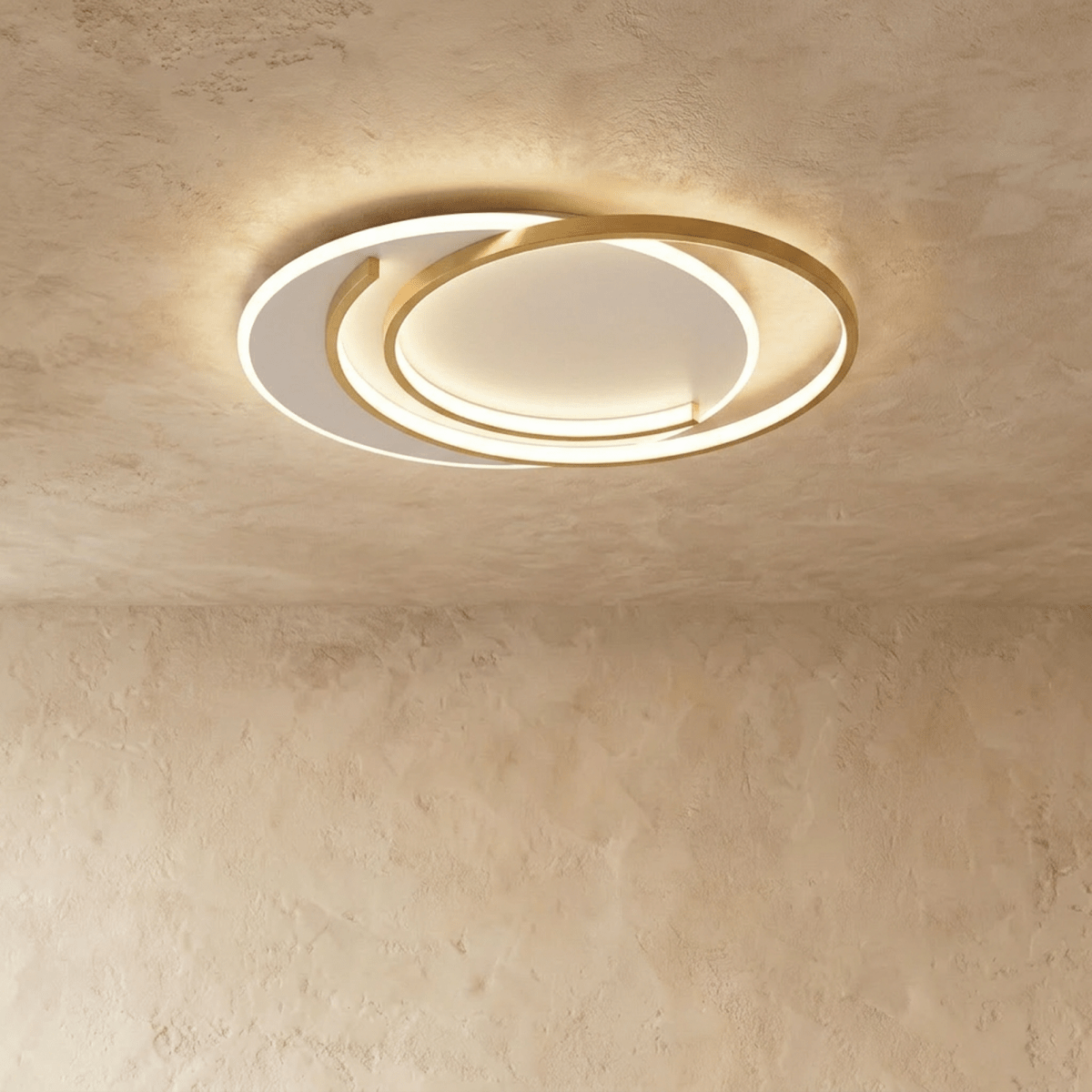 Clérachic flush ceiling light of gold aluminium rings in a crescent layout, glowing warm on a limewash ceiling.