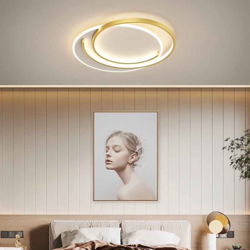 Clérachic gold crescent-ring ceiling light glowing above a bed, framed portrait and panelled wall in a calm bedroom.