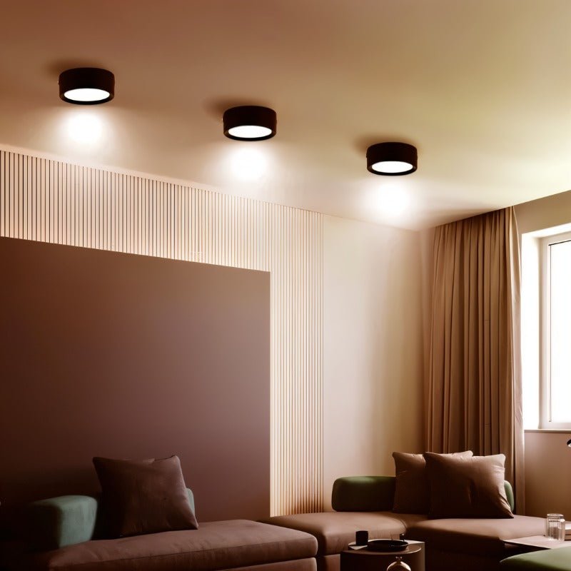 Clérachic three black round surface-mounted downlights across the ceiling of a warm living room with a corner sofa and curtains.