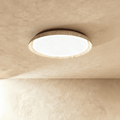 Clérachic flat round ceiling light with a travertine-look ring and white diffuser, mounted on a textured sand-toned plaster ceiling.