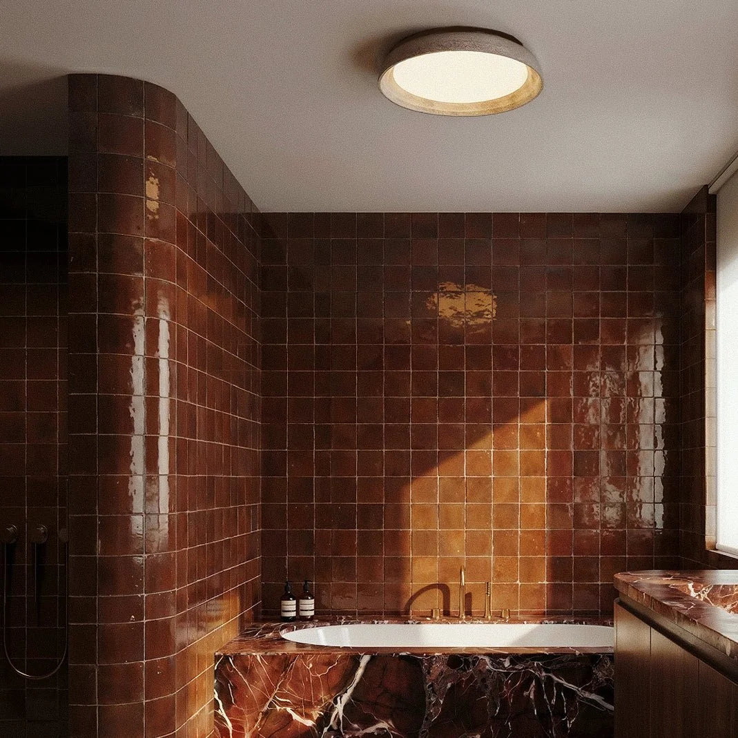 Clérachic flush ceiling light with a pale stone-look rim and warm glowing diffuser, above a tiled room with a marble-clad tub.