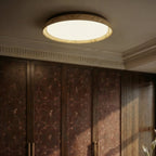 Clérachic flat ceiling light with a veined stone-look ring and opal diffuser, glowing above a cornice and embroidered wooden wardrobe doors.