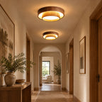 Two Clérachic wood-grain ceiling lights glowing along a warm hallway with an arched doorway, a stone vase of foliage and a wooden console.