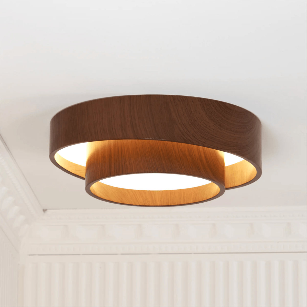 A Clérachic ceiling light glowing amber between its stacked rings, beside a white cornice and fluted wall.