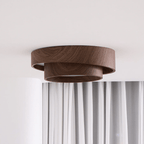 Side view of a Clérachic ceiling light with two stepped wood-grain rings, hung over white curtains on a pale wall.