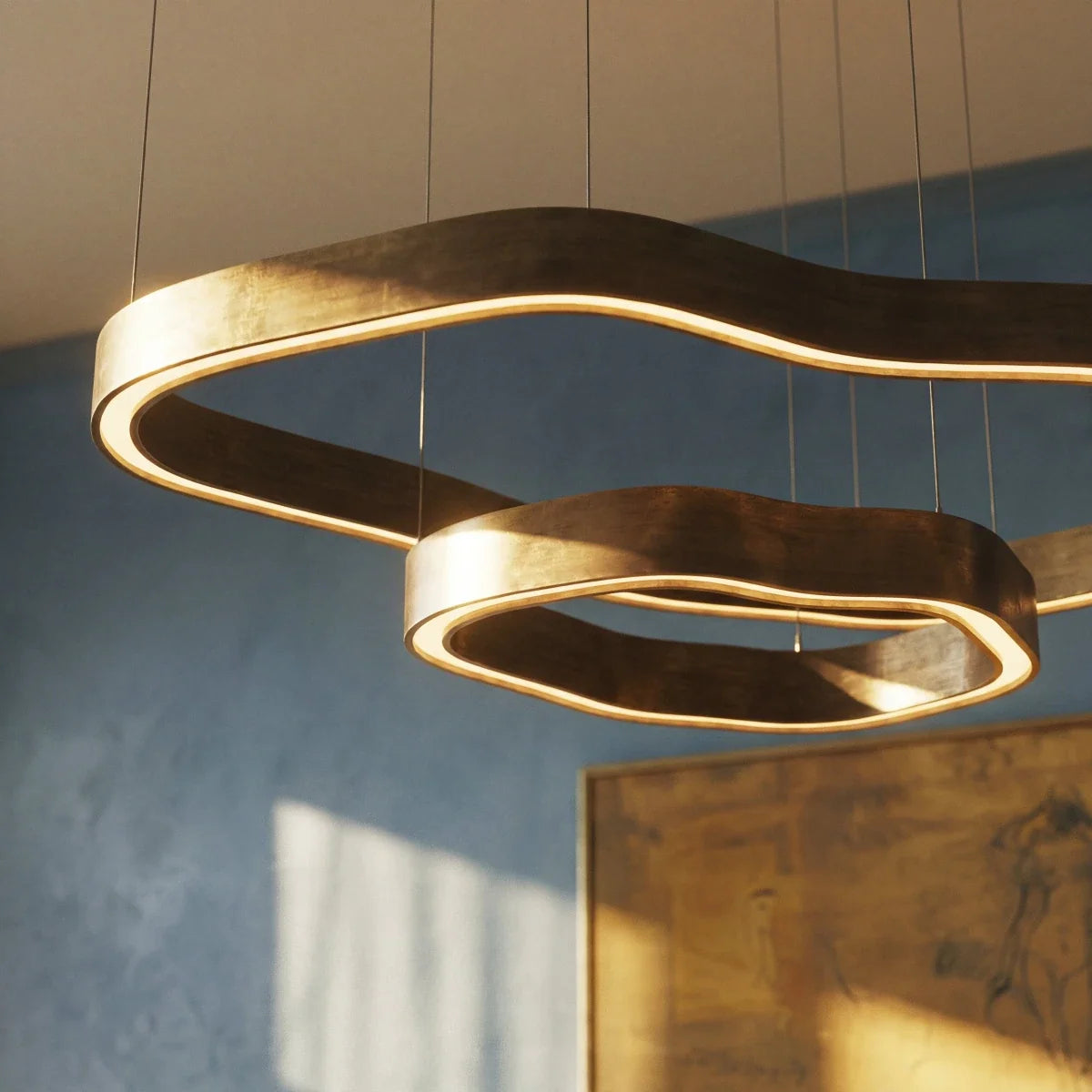 Close view of two wavy Clérachic chandelier rings in aged brass, their LED edge glowing warmly against a blue plaster wall.