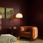 Clérachic floor lamp with a white glass dome glowing beside a rust bouclé armchair in a deep burgundy sitting room.