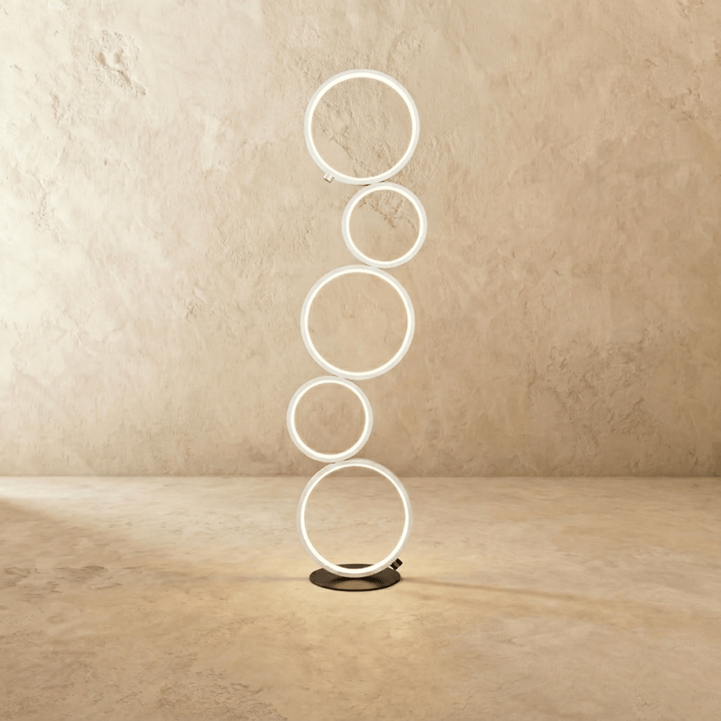 Clérachic floor lamp of five stacked white LED rings lit warm white on a dark round base, studio shot on a pale limewash wall.
