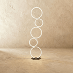 Clérachic floor lamp of five stacked white LED rings lit warm white on a dark round base, studio shot on a pale limewash wall.