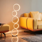 Clérachic stacked-ring LED floor lamp glowing beside a mustard leather sofa and footstool in a warm living room.