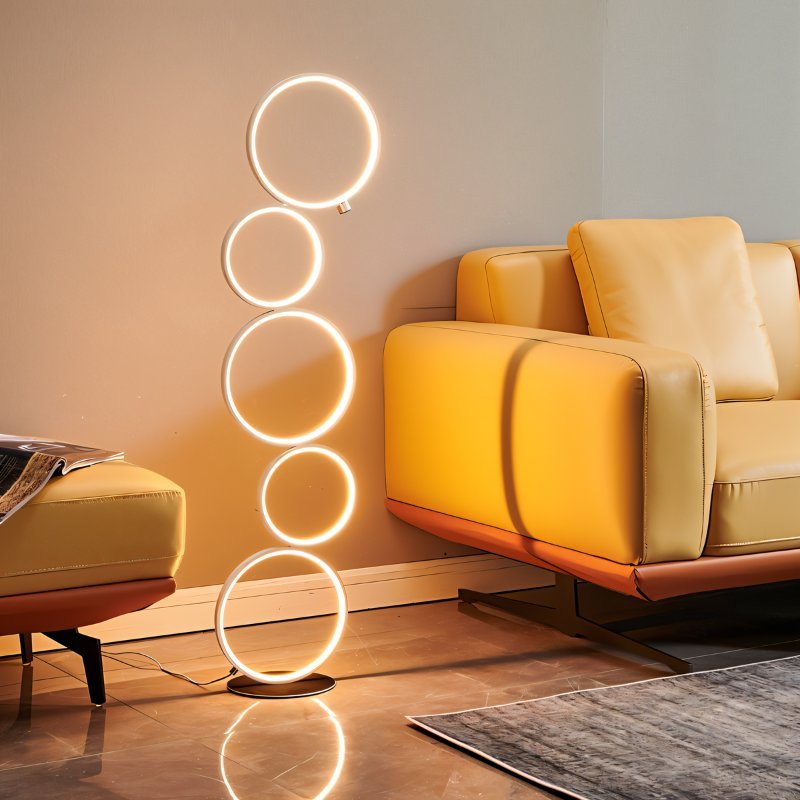 Clérachic stacked-ring LED floor lamp glowing beside a mustard leather sofa and footstool in a warm living room.
