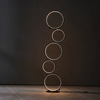 Clérachic stacked-ring floor lamp in black with warm white light, standing in a dark room against a charcoal wall.