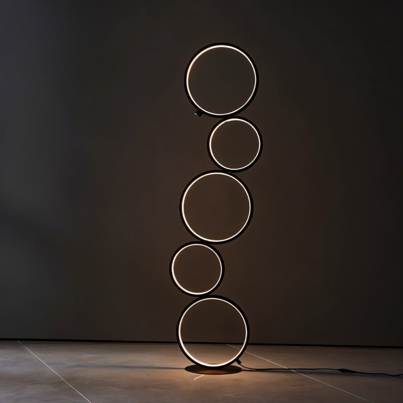 Clérachic stacked-ring floor lamp in black with warm white light, standing in a dark room against a charcoal wall.