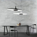 A black disc pendant and a smaller white one hang at different heights over a long black table in front of a grey stone-effect wall, from Clérachic.