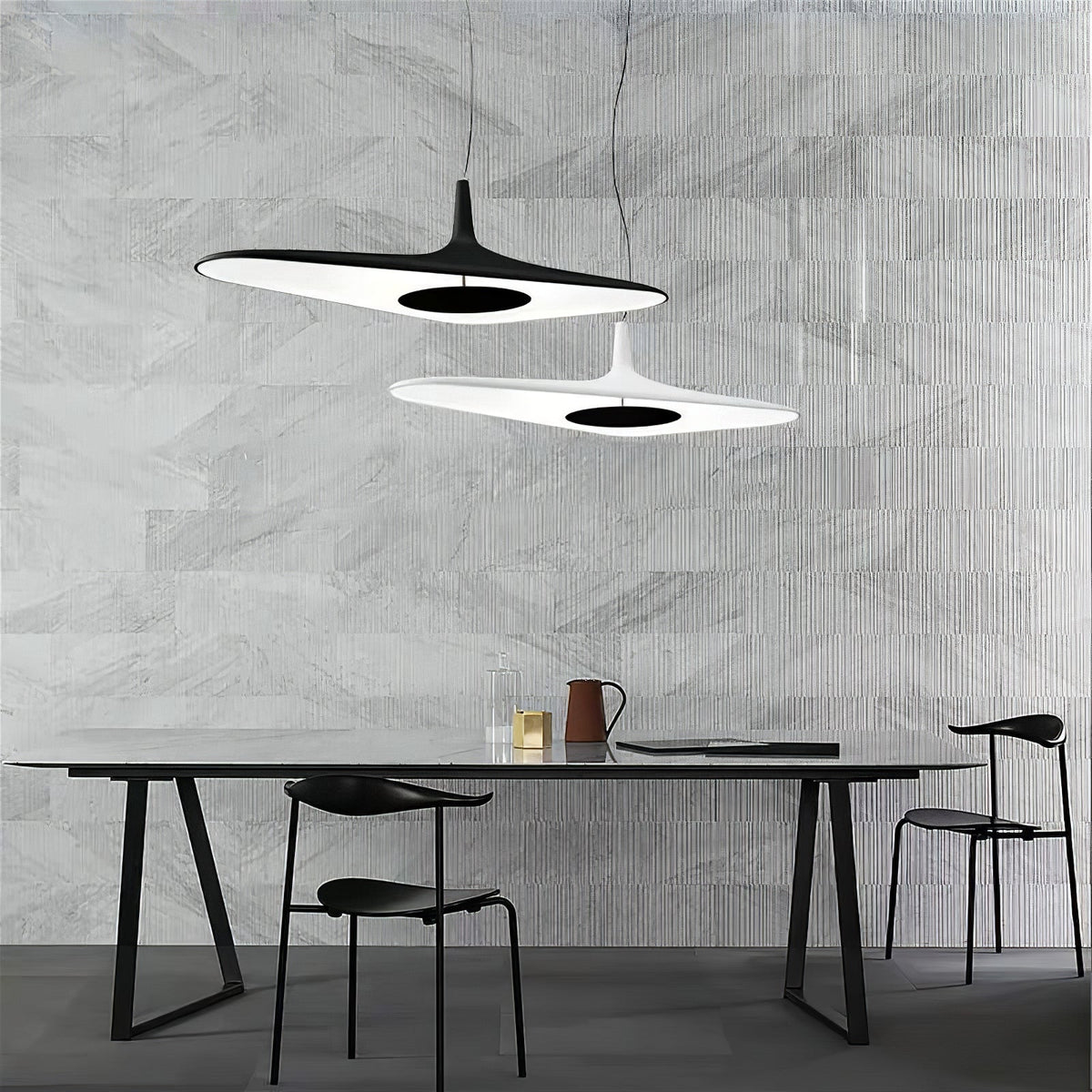 A black disc pendant and a smaller white one hang at different heights over a long black table in front of a grey stone-effect wall, from Clérachic.