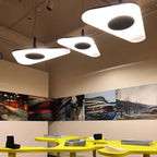 Three triangular-edged white disc pendants glow at staggered heights in a gallery-style room with a yellow table, shown as this Clérachic LED pendant.