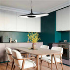 A black disc pendant hangs low over a wooden dining table with cream chairs, in front of a deep teal kitchen wall, a Clérachic LED pendant.