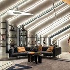 A white disc pendant hangs below a slanted ceiling with strip lighting, above a dark sofa and a display wall of figures, a Clérachic LED pendant.