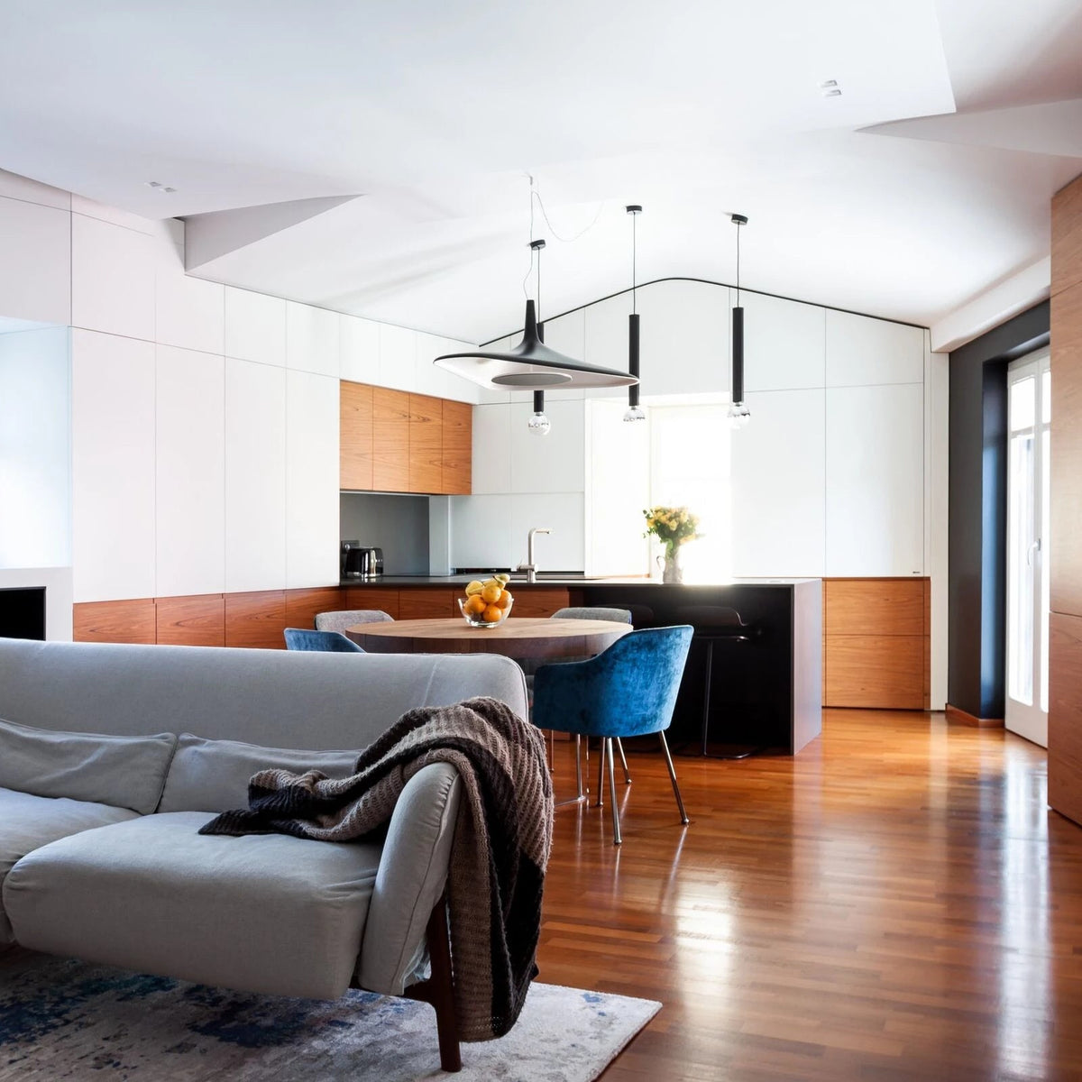 A black disc pendant hangs in an open living and kitchen space with a grey sofa and blue chairs, beside slim black pendants, from Clérachic.