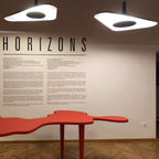Two white disc pendants glow overhead beside a wall of printed text, with a red sculptural table below, in a Clérachic LED pendant setting.